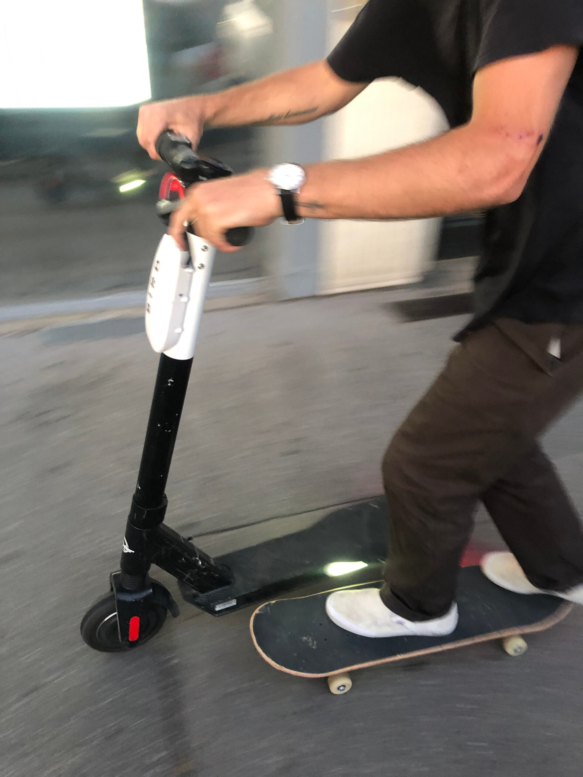 Ein Mann mit Skateboard hat seine Hände am Lenker eines Elektrorollers und lässt sich damit die Straße entlang ziehen.